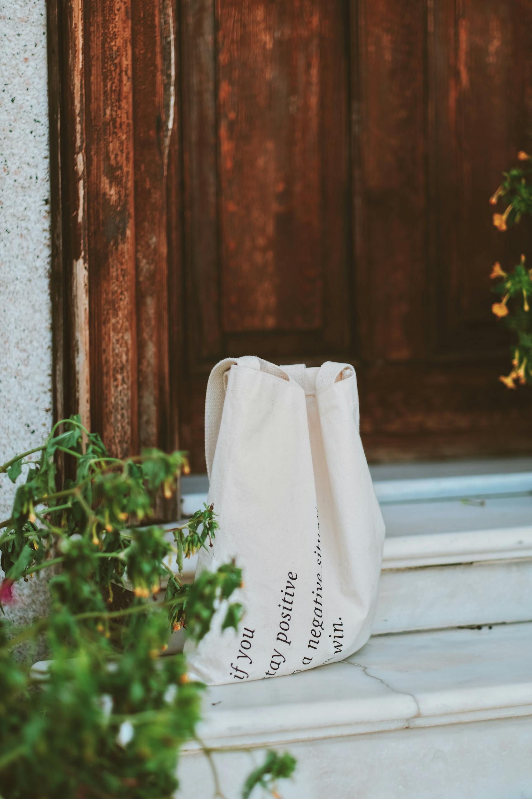 A white tote bag sits on marble steps in front of a rustic wooden door surrounded by green plants.