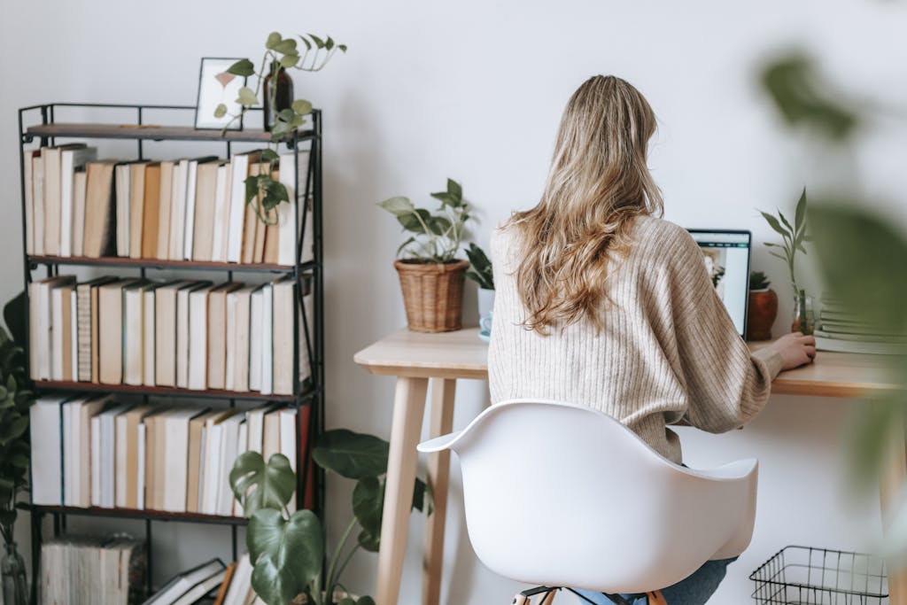 Adult woman working on a laptop at a home office desk surrounded by books and plants.