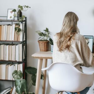 Adult woman working on a laptop at a home office desk surrounded by books and plants.