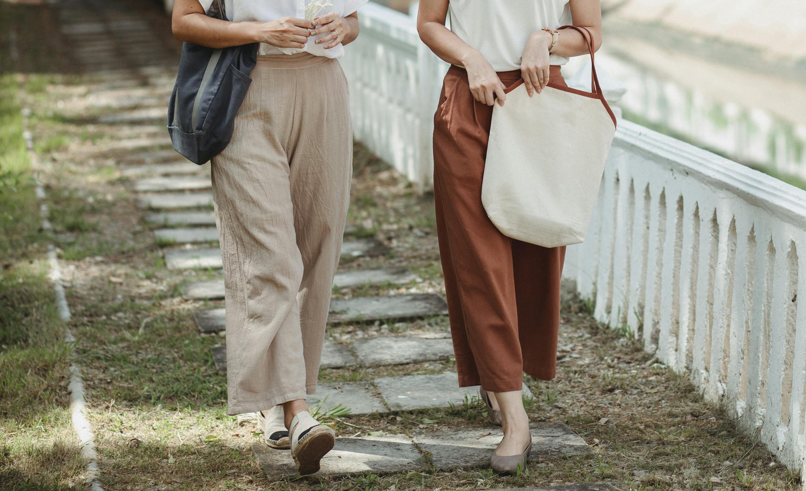 Crop unrecognizable girlfriends in casual wear with bags strolling on fenced walkway in town in summer